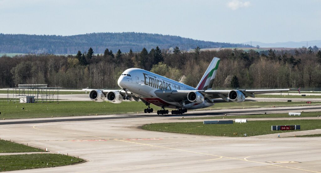 NRI & Global Services Emirates Airbus A380 taking off in daylight at Zurich Airport with scenic backdrop.
